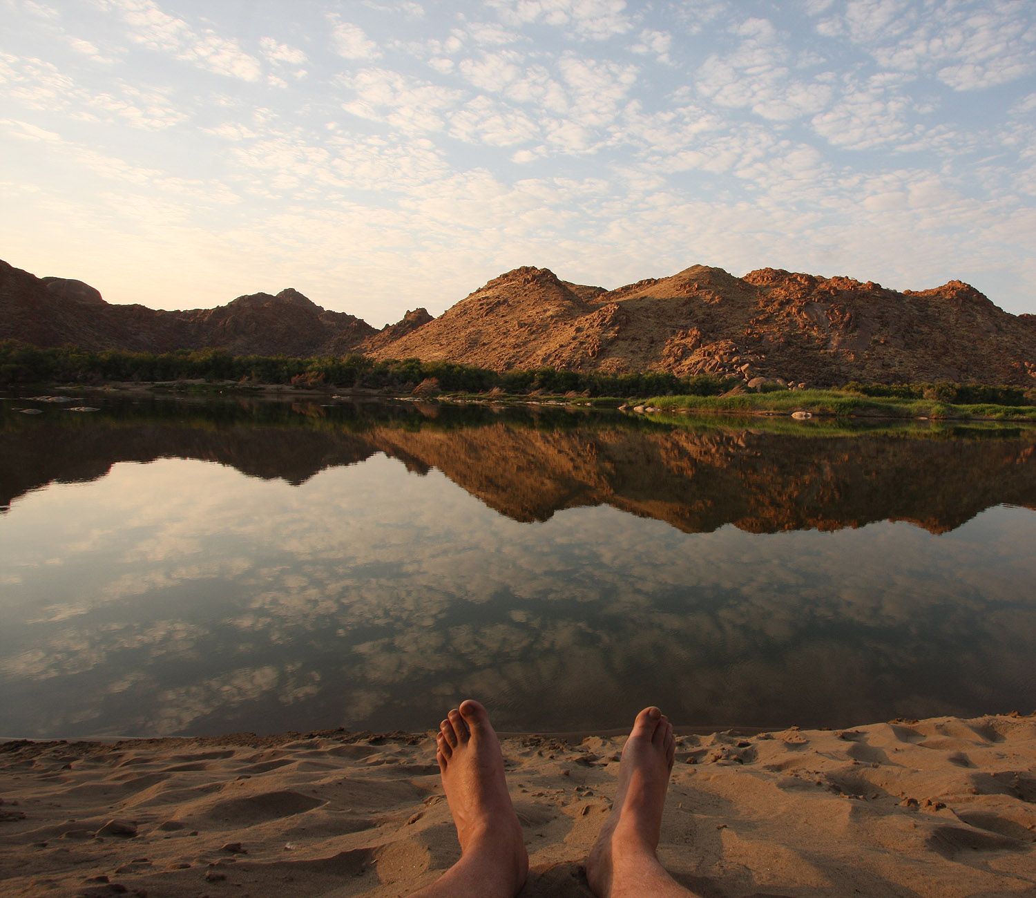 Looking out from a riverbank at dusk across calm water and hills.