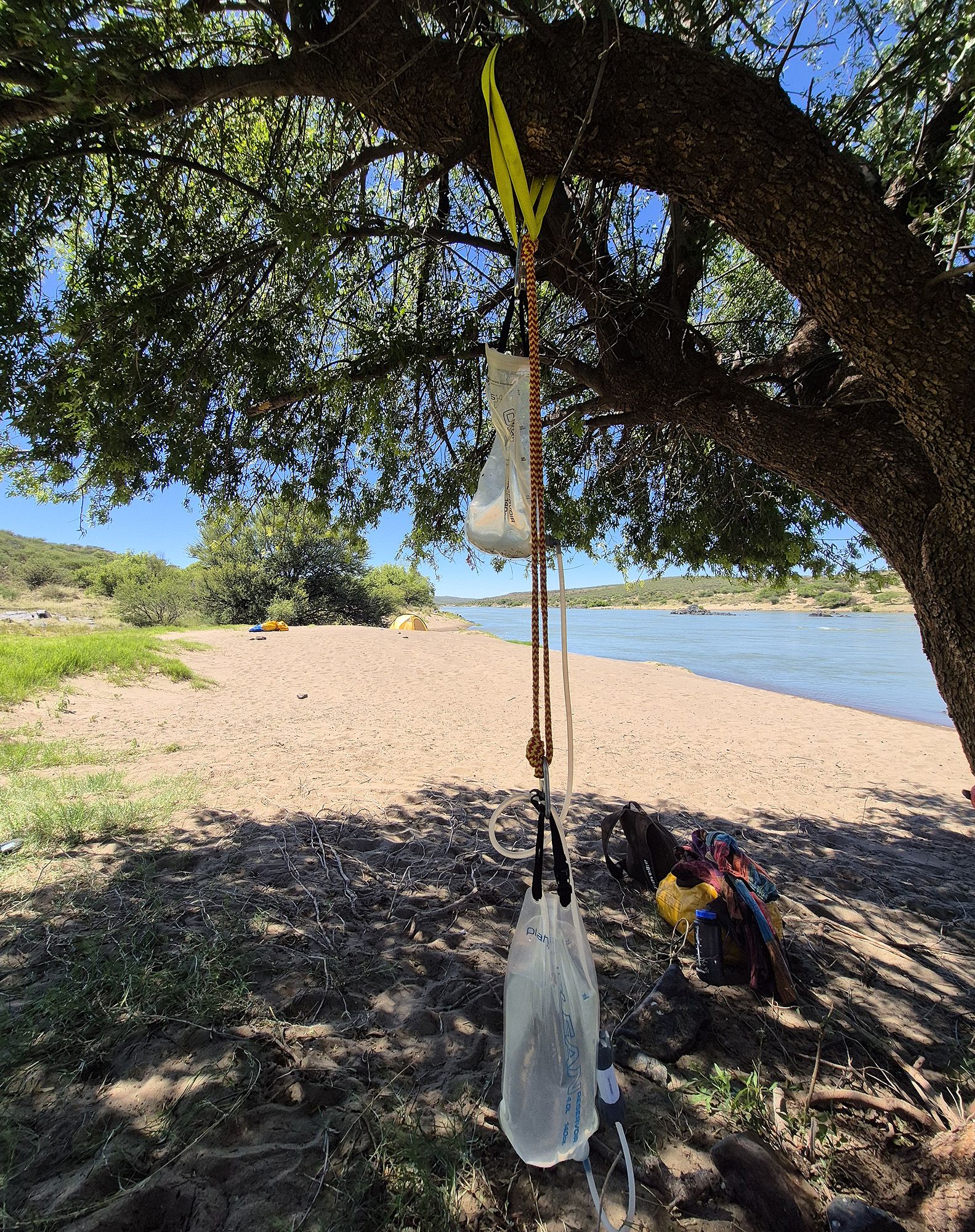 A gravity water filter hanging from a tree at a riverside campsite.