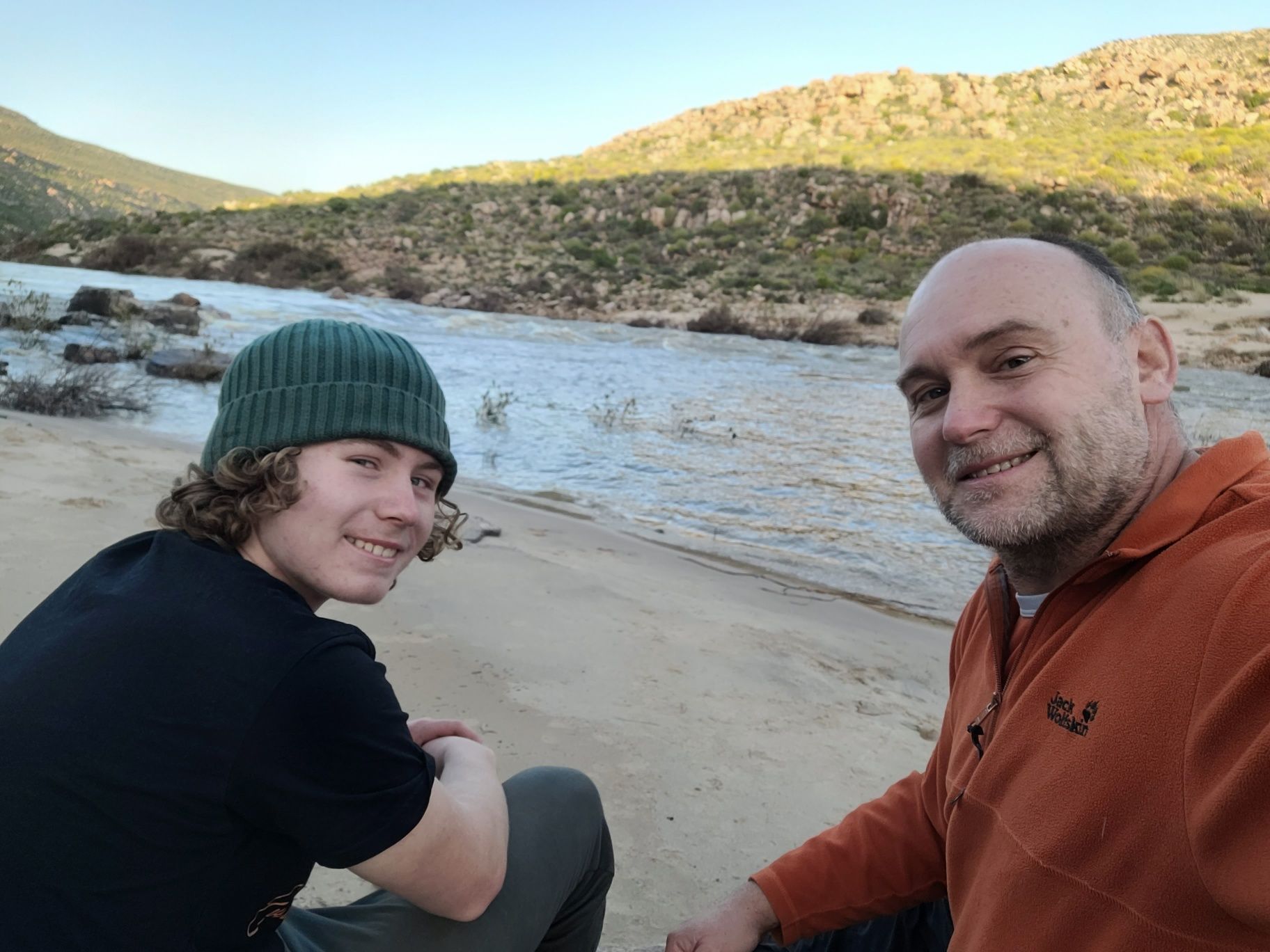 Celliers and Ruben sitting on a sandy riverbank, smiling, with the river and hills behind them.