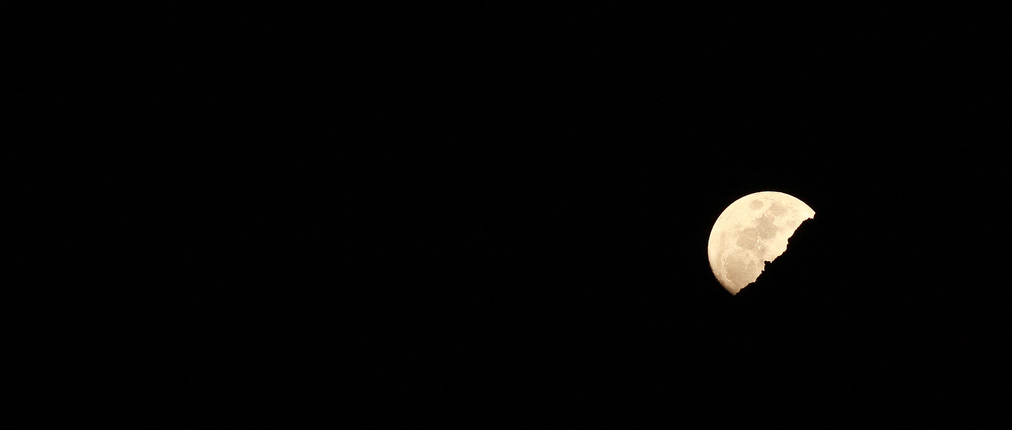 A pale moon rising behind a dark ridgeline in a black night sky.